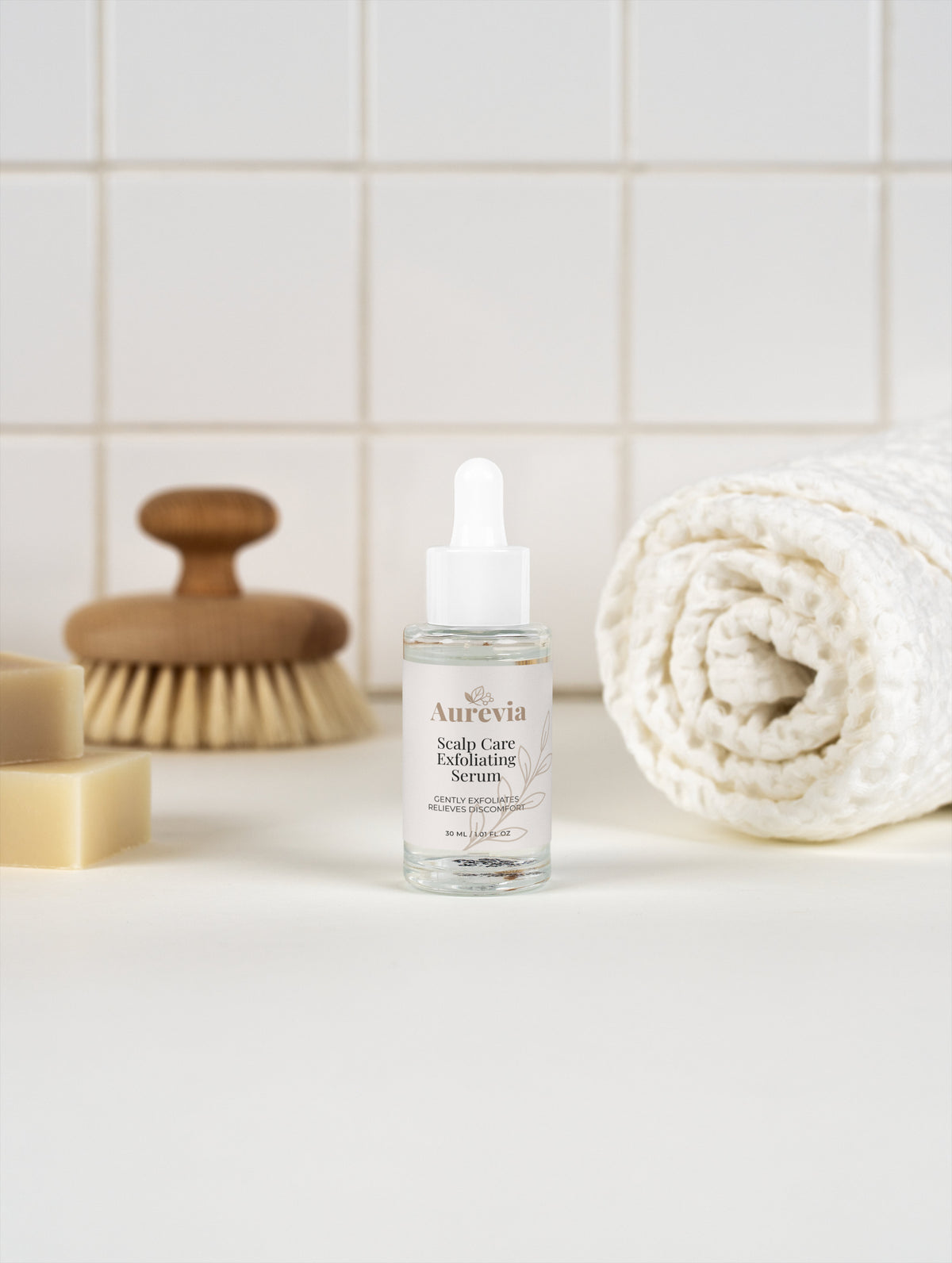 Aurevia Scalp Care Exfoliating Serum bottle with dropper, beside wooden brush and towel on tiled bathroom counter