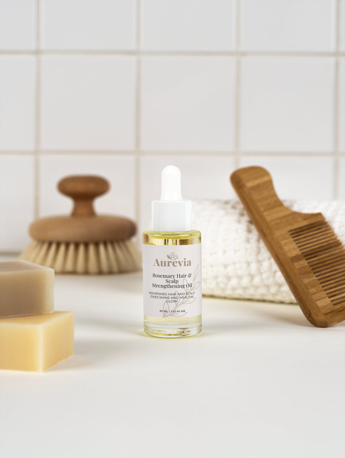 Rosemary Hair & Scalp Strengthening Oil with wooden comb and soap bars on tiled background